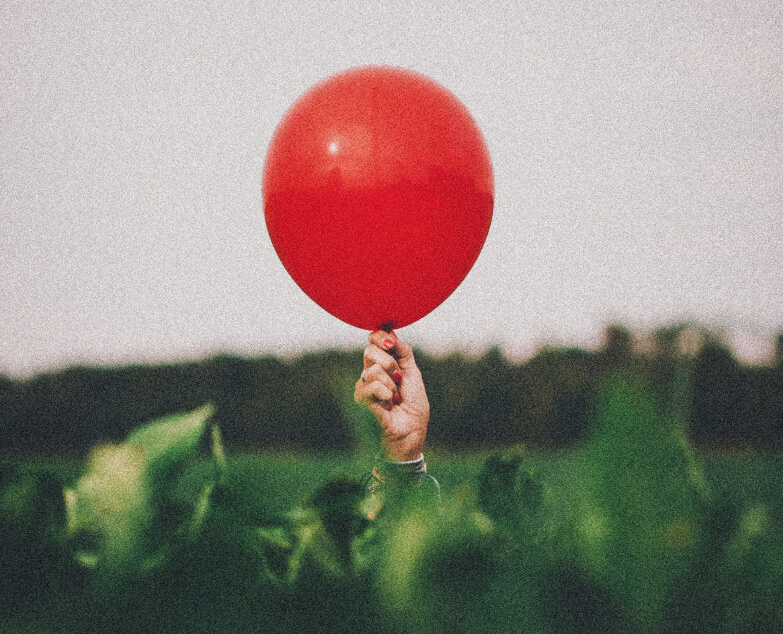 A 90s style photograph of a hand reaching out from a bush holding an inflated red balloon.