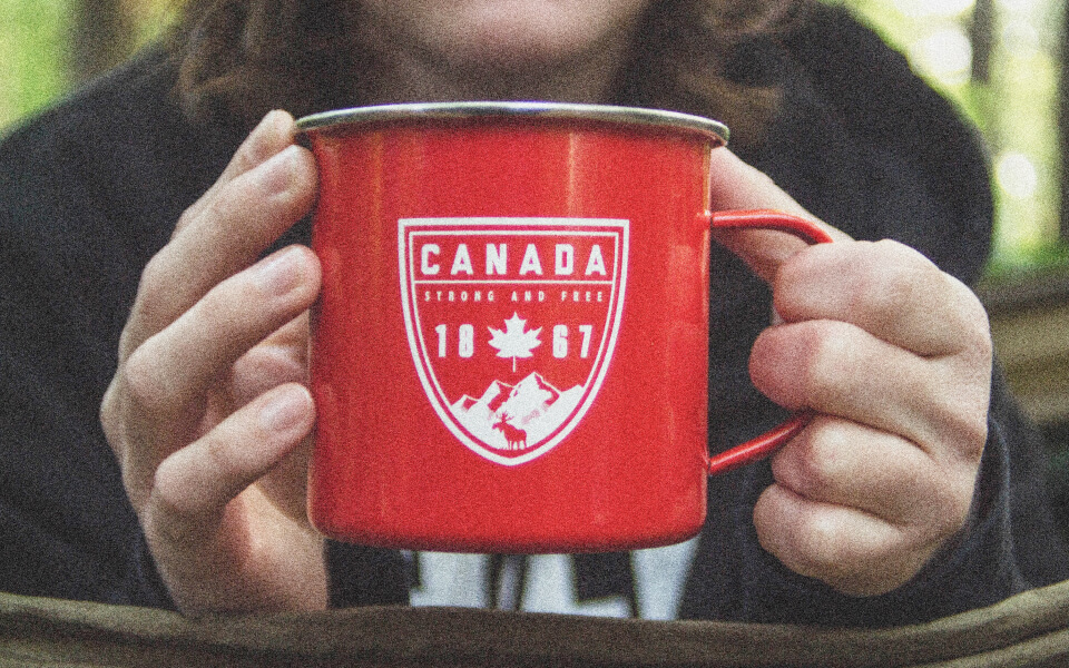 A 90s style photograph close-up of someone in the Canadian forest holding a red camping mug reading "Canada, Strong and Free, 1867".